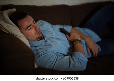 Portrait Of Young Man Falling Asleep On Couch In Dark Room , Holding Remote Controller While Watching TV Late At Night