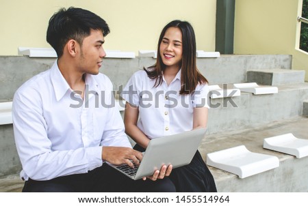 Portrait of young man and girl in university uniform sitting and working on laptop. Nice Asia girl sitting with friend and holding laptop in hands. Boy and girl happily looking at laptop
