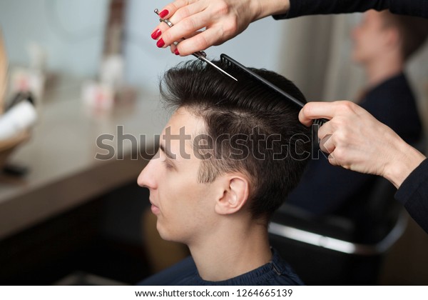 portrait of young man having\
haircut in barber shop. Hairdresser cutting man\'s hair with\
scissors