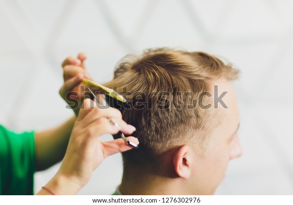 portrait of young man having\
haircut in barber shop. Hairdresser cutting man\'s hair with\
scissors.