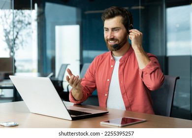 Portrait of a young man in a headset and a red shirt. He sits at the desk in the office, works on a laptop, communicates through a video call, talks, smiles. - Powered by Shutterstock