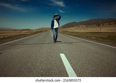 Portrait Of A Young Man Hitchhiker In Denim Clothes And Black Sunglasses Walking On The Highway On The Background Of Picturesque Landscape. Denim Fashion. Road Adventures, Travelling.