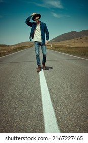 Portrait Of A Young Man Hitchhiker In Denim Clothes, Black Sunglasses And Hat Walking On The Highway On The Background Of Picturesque Landscape. Denim Fashion. Road Adventures, Travelling.