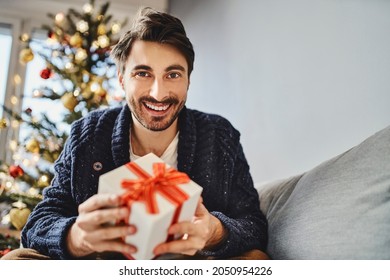 Portrait Of Young Man Holding Christmas Gift Sitting On The Sofa At Home