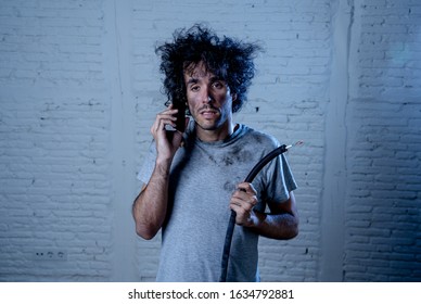 Portrait Of Young Man Holding Electrical Cable After Domestic Accident With Dirty Burnt Funny Face Expression Calling Desperate With Mobile Phone Asking For Help. Electricity Repairs And DIY Concept.