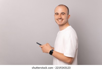 Portrait Of Young Man Holding Smartphone And Smiling Looking At The Camera Over Grey Backdrop.