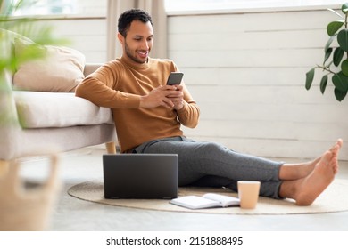 Portrait Of Young Man Holding Using Cell Phone Sitting On Floor Leaning On Couch In Living Room. Cool Guy Browsing Internet, Surfing Web Watching Video, Using Application And Pc, Full Body Length