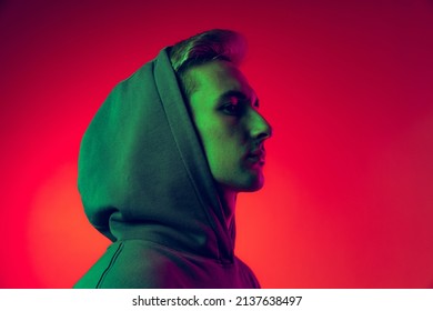 Portrait of young man in hood looking away, posing isolated over red studio background in green neon light. Fashionable youth. Concept of facial expression, fashion, youth, lifestyle, emotions
