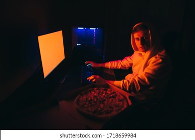 Portrait Of Young Man In Hoodie Sitting At Computer At Home Near Pizza Box At Night, Looking Into Camera With Serious Face. Freelancer Guy Works At His Computer At Night And Eats Fast Delivery Food.