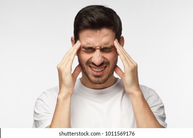 Portrait of young man isolated on grey background suffering from severe headache, pressing fingers to temples, closing eyes to relieve pain with helpless face expression