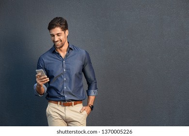 Portrait Of Young Man Leaning Against A Grey Wall Using Mobile Phone. Happy Business Man Messaging With Smartphone Isolated On Gray Background With Copy Space. Smiling Guy Typing And Reading A Message