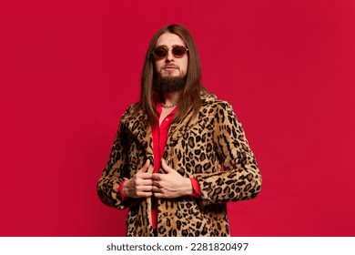 Portrait of young man with long hair posing in stylish fur coat and sunglasses against red studio background. Charismatic look. Concept of emotions, facial expression, lifestyle, unique style