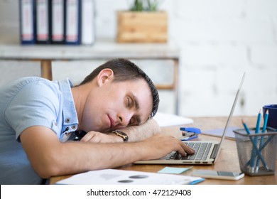 Portrait of young man lying on the table in front of laptop, sleepy, tired, overworked or lazy to work. Attractive business man napping in his home office relaxing after work on laptop computer