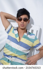 Portrait Of Young Man Model In Sunglasses, Summer Polo Shirt Sitting Chair With Green Palm Shadow Posing In Studio