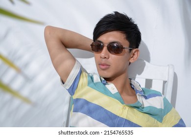 Portrait Of Young Man Model In Sunglasses, Summer Polo Shirt Sitting Chair With Green Palm Shadow Posing In Studio