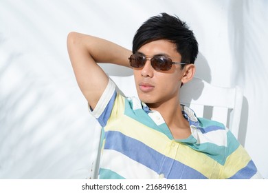 Portrait Of Young Man Model In Sunglasses, Summer Polo Shirt Sitting Chair With Green Palm Shadow Posing In Studio