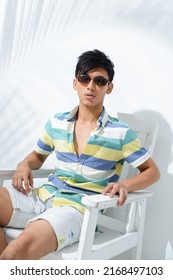 Portrait Of Young Man Model In Sunglasses, Summer Polo Shirt Sitting Chair With Green Palm Shadow Posing In Studio