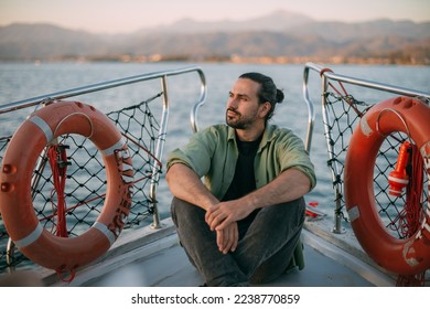 Portrait of a young man on a boat trip on a small ship in the sea on a sunny day. A Caucasian tourist guy in a summer shirt on the deck of a boat enjoys the wind in his face at sunset - Shutterstock ID 2238770859