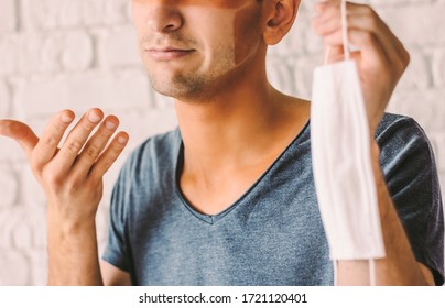 Portrait Of Young Man Patient With Funny Sun Tanned Lines On Face After Wearing Medical Face Mask. Confused Hipster Man With Sunburn Tan Face Skin Holding Protective Mask In Hand. COVID-19 Quarantine