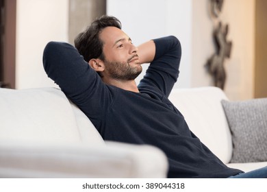 Portrait Of A Young Man Resting On Sofa And Thinking About The Future. Handsome Young Man With Hands Behind Head Sitting On Couch In Living Room. Positive Man Daydreaming And Relaxing At Home. 
