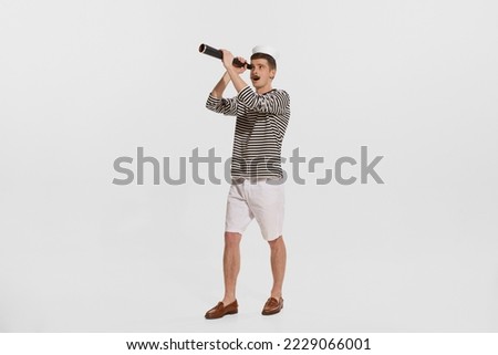 Portrait of young man, sailor in striped shirt looking in spyglass isolated on white background. Curious person. Concept of summer holidays, occupation, retro fashion, vintage style. Copy space for ad