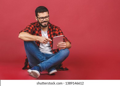 Portrait of young man sitting on the floor using a tablet pc, isolated against red background. Ready for your design.