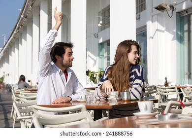 Portrait of a young man sitting at outdoor cafe with raised arm asking for waiter