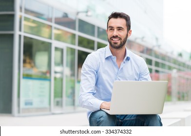 Portrait Of Young Man Sitting Outside With Laptop. He's Looking Away. Shallow Depth Of Field With Focus On Young Man Sitting With Laptop.
