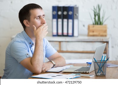 Portrait of young man sitting at the table in front of laptop, sleepy, tired, overworked or lazy to work. Attractive business man yawning in home office relaxing or bored after work on laptop computer