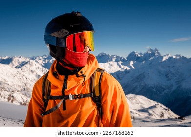 Portrait of a young man in a ski mask, stands in a ski resort against the backdrop of mountains and blue sky