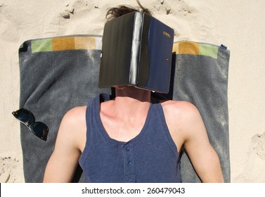 Portrait Of Young Man Sleeping On Beach With Book Covering Face