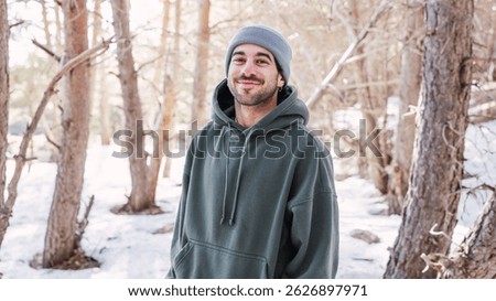 Portrait of a young man smiling while enjoying a snowy forest landscape, dressed in a cozy hoodie and warm beanie, embracing winter's chill