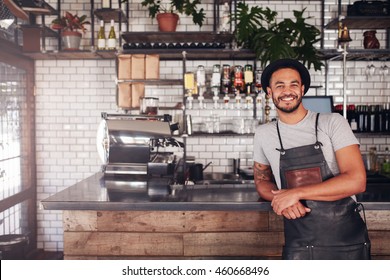 Portrait of young man standing at the counter in his cafe. Coffee shop working in apron and hat smiling at camera. - Powered by Shutterstock