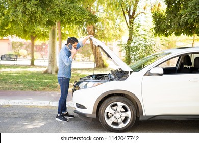Portrait of young man standing in front of his broken car, looking at car engine and talking on cell phone