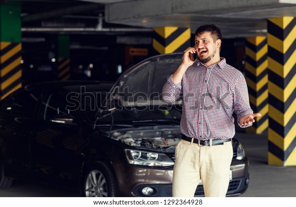 Portrait of a young man\
standing in front of his broken car and talking on his cell phone\
calls for help