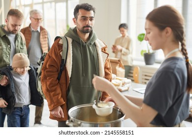 Portrait Of Young Man Standing In Line At Soup Kitchen With Young Woman Giving Out Simple Meals To People In Need