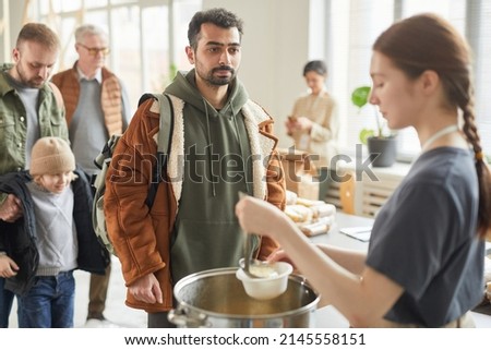 Portrait of young man standing in line at soup kitchen with young woman giving out simple meals to people in need