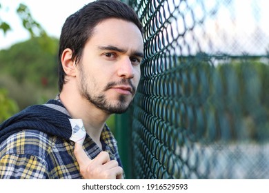 Portrait of young man standing next to a fence holding his jacket over his shoulder watching sports. Guy looking behind the camera.