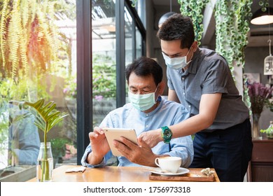 Portrait of a young man teaching his father how to use tablet computer and wearing mask for prevent the spread of the Covid-19. Father and Son using a tablet together at cafe.