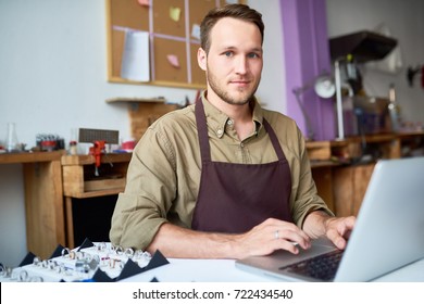 Portrait of young man using computer cataloging jewelry while appraising goods in pawn shop and looking at camera
