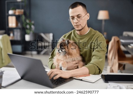 Portrait of young man using computer and holding cute dog in lap while working in pet friendly office