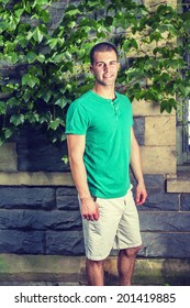 Portrait Of Young Man. Wearing A Green Short Sleeve Henley Shirt, Light Yellow Shorts, A Young Guy Is Standing By The Wall With Green Ivy Leaves, Smiling, Looking At You. 