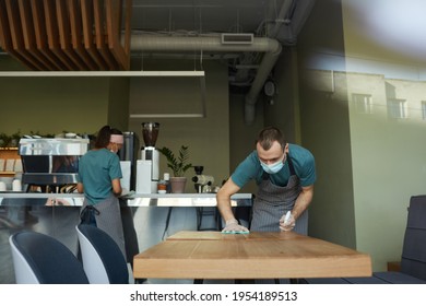 Portrait Of Young Man Wearing Mask While Cleaning Tables With Sanitizer At Cafe Or Coffee Shop, Covid Safety Measures, Copy Space