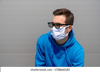 Portrait of a young man in a white mask and black glasses, dressed in a blue hoodie with an earring, with a serious face look into the distance, standing against a gray textured wall in the afternoon - Powered by Shutterstock