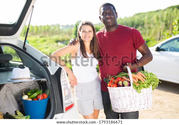 Portrait of young man and\
woman with basket full of freshly picked vegetables standing near\
car