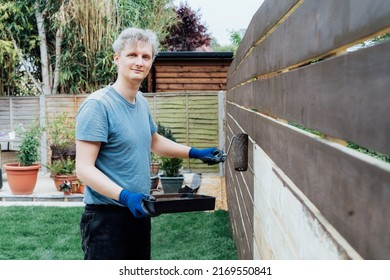 Portrait Of Young Man Worker Paints With A Roller A Wooden Board Fence In The Garden. DIY, Do It Yourself Concept. House Improvement. Home Renovation And Refurbishment. Selective Focus, Copy Space.