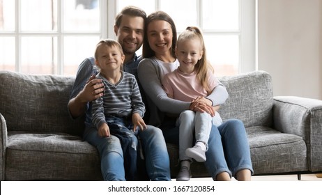 Portrait Of Young Married Couple Resting On Sofa, Holding On Lap Adorable Kids Siblings. Happy Loving Bonding Family Of Four Relaxing On Couch, Posing For Photo Together In Modern Living Room.