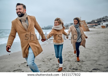Portrait of a young married couple and their cute daughter who have fun on the beach in winter wearing warm clothes and scarves in the cold season.