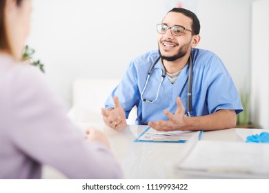 Portrait of young Middle-Eastern doctor wearing glasses talking to female patient sitting at desk in office and gesturing actively - Shutterstock ID 1119993422