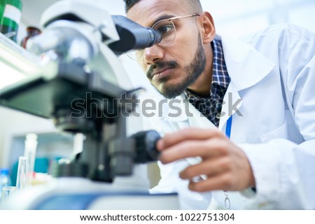Portrait of young Middle-Eastern scientist looking in microscope while working on medical research in science laboratory, copy space
